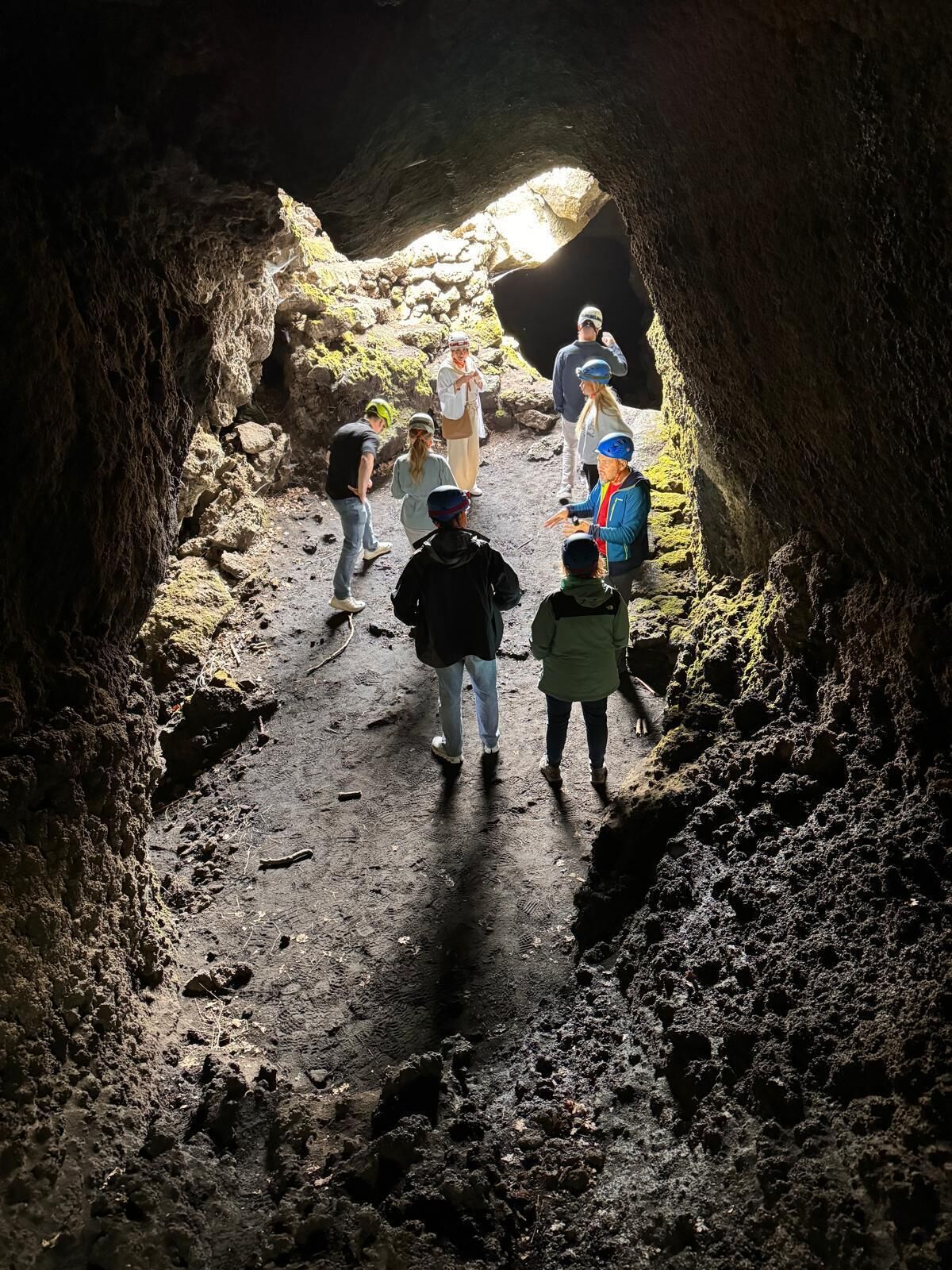 A group of people walking through a narrow cave passage with rough, rocky walls and a dirt floor.