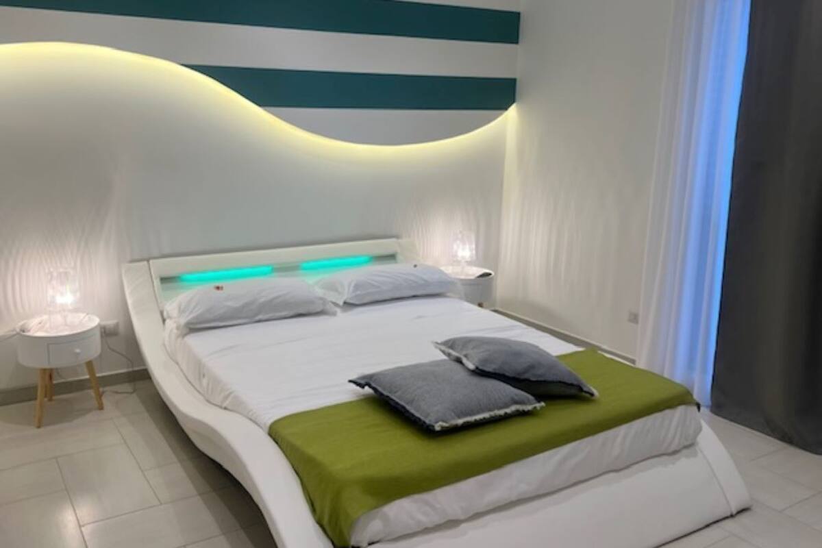 A modern bedroom with a curved headboard, a bed with white bedding and green accents, two bedside lamps, and a view of a window with curtains.