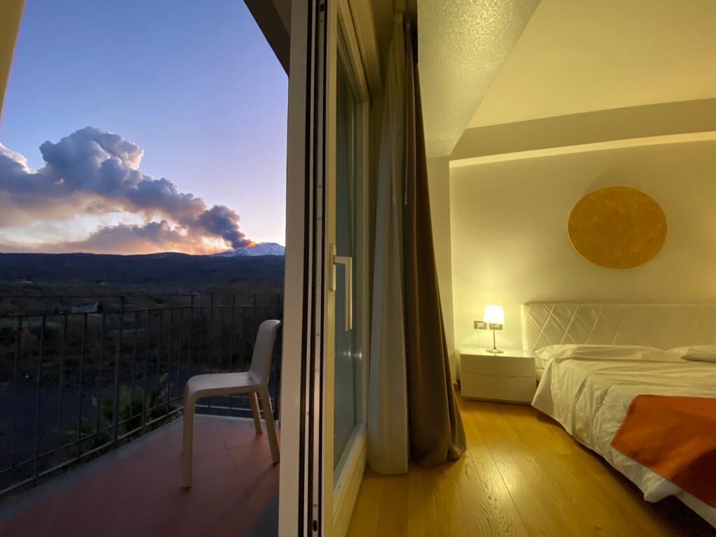 A bedroom with a bed, a bedside table with a lamp, and a view of the cityscape through a balcony.