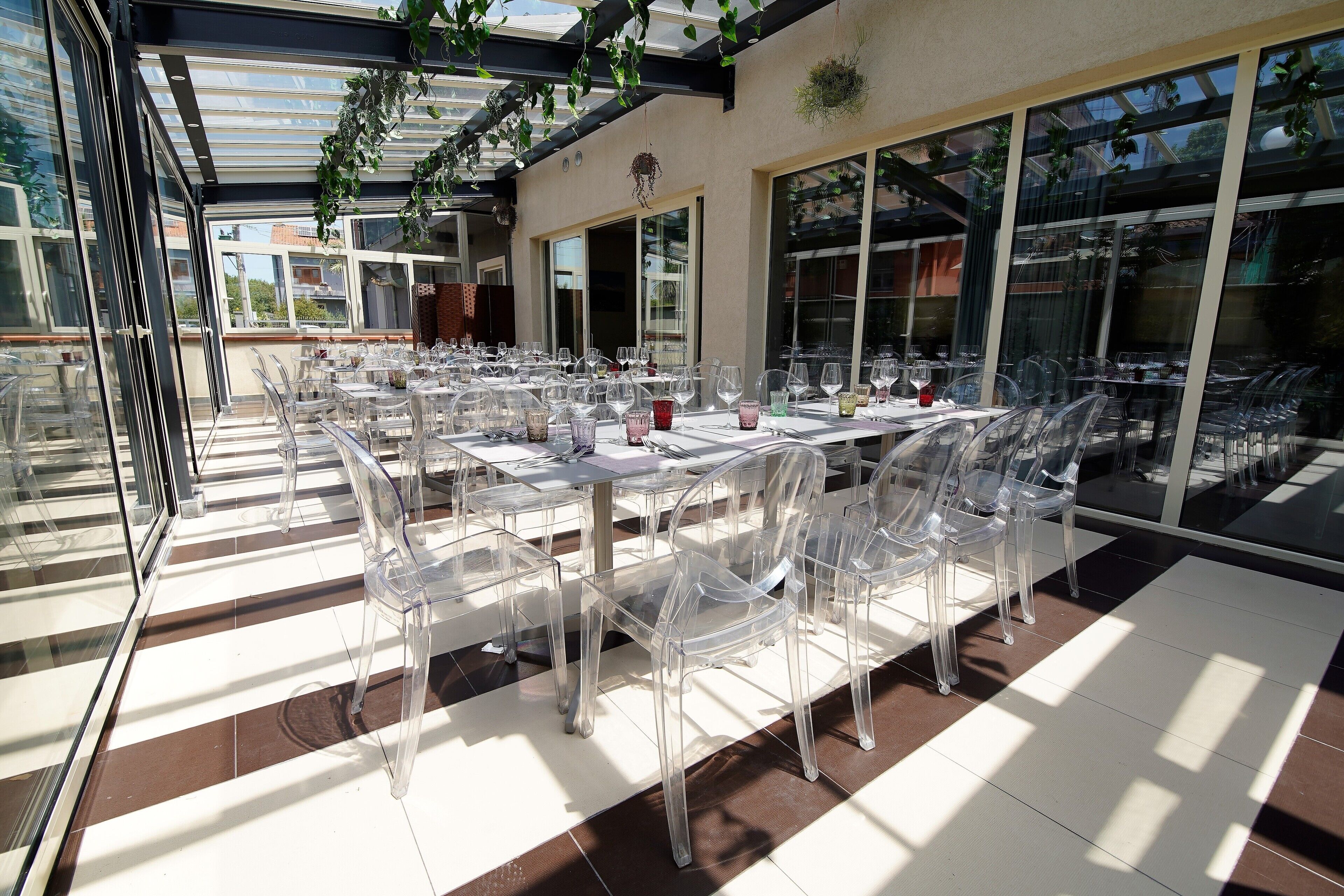 Un espace de restauration en plein air avec des chaises et des tables en plastique transparent, de grandes fenêtres et une structure en forme de pergola.
