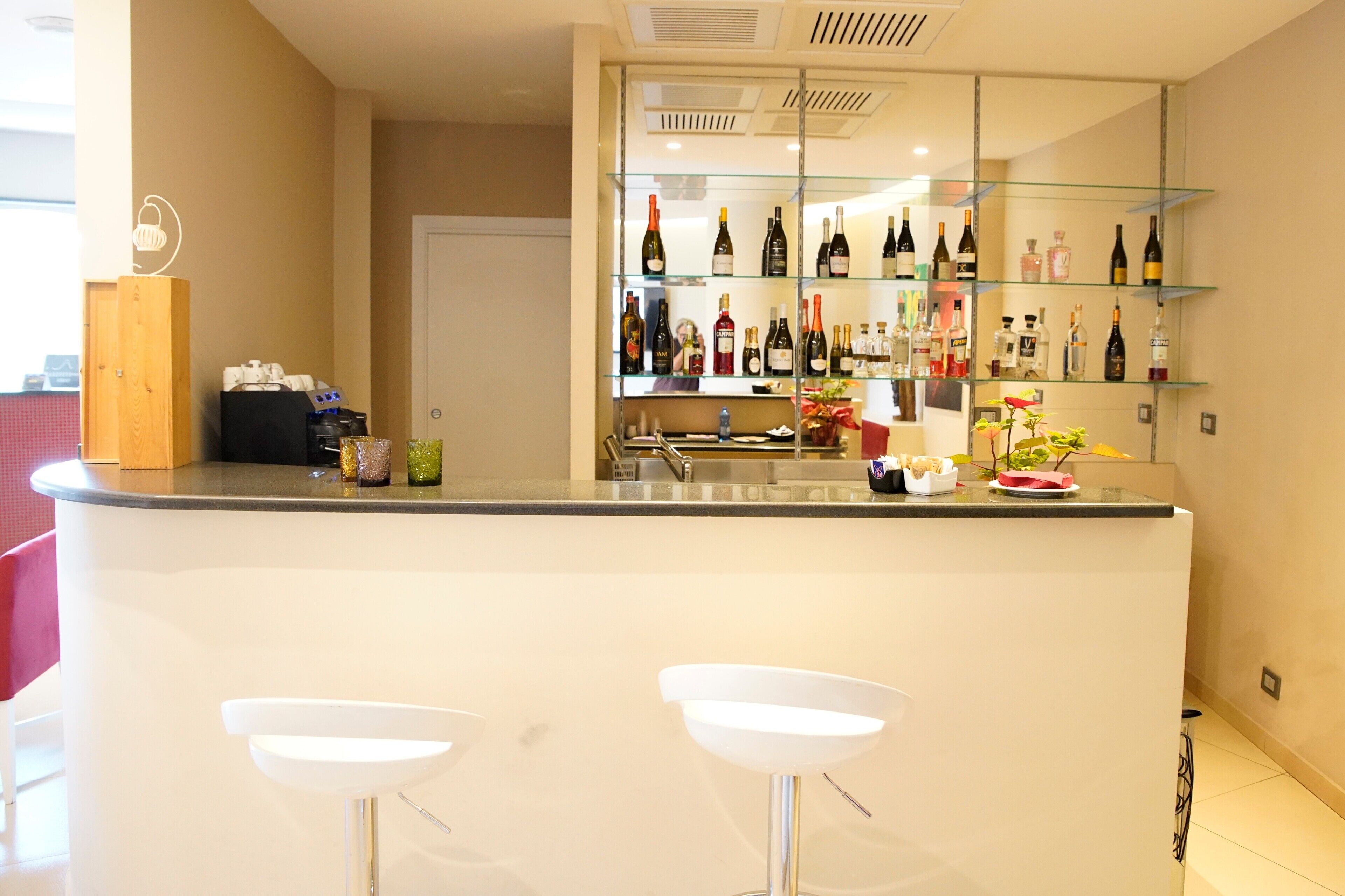 A modern bar area with a glass shelf displaying various bottles, two white bar stools, and a coffee machine.