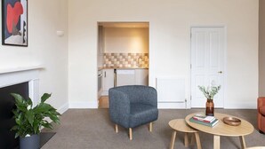 Una sala de estar moderna con un sillón gris, una mesa de centro redonda de madera y una estantería. Se ve una chimenea a la izquierda y una cocina al fondo.