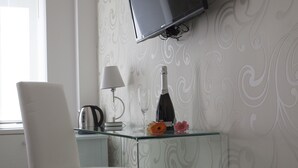 A modern hotel room with a glass desk, a white chair, a flat-screen TV mounted on the wall, a lamp, a bottle of champagne, and a trash bin. - Elenoire Rooms & Suite (Rome)