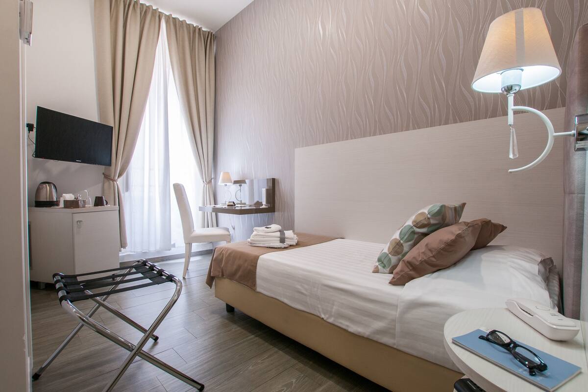 A hotel room with a bed, bedside table, lamp, TV, desk, chair, and a drying rack.