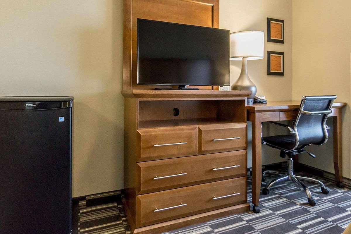 A hotel room with a wooden TV stand, a flat-screen TV, a desk with a chair, and a refrigerator.