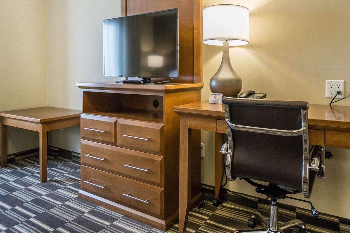 A hotel room with a flat-screen TV, a desk with a lamp, a chair, and a wooden dresser with drawers.