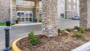 Fachada de hotel con pilares de piedra, puerta de cristal y zona de estacionamiento.