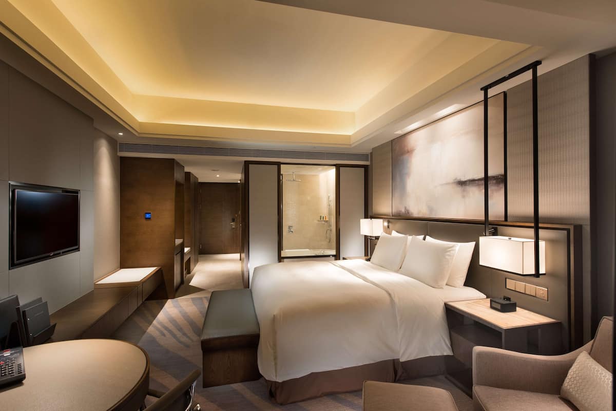 A modern hotel room with a large bed, bedside tables, a desk, a TV, and a shower area.