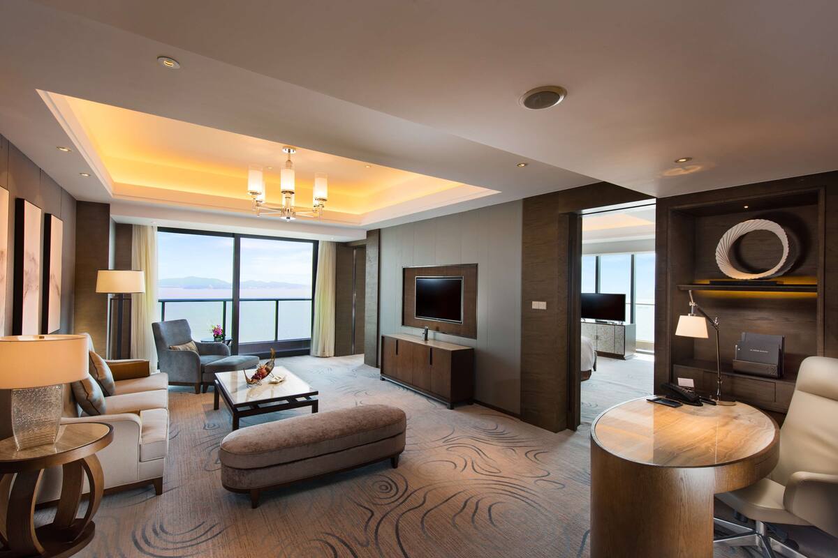 A modern hotel room with a large window offering a view of the sea, a flat-screen TV mounted on the wall, a round coffee table, and a comfortable seating area with a sofa and armchairs.