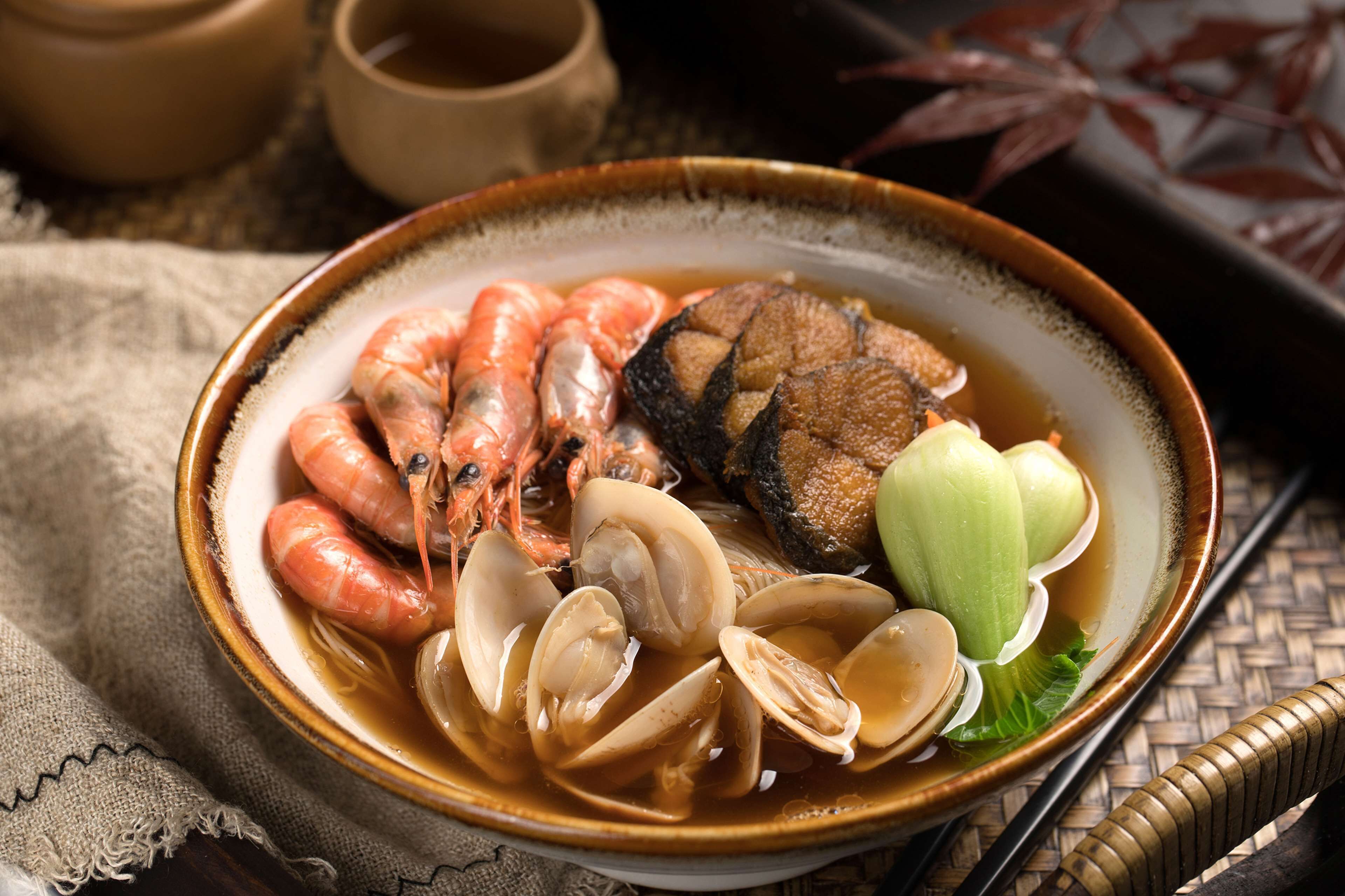 Una ciotola di zuppa di pesce con gamberi, vongole e pesce, servita in una ciotola di ceramica su un sottotavola intrecciato.