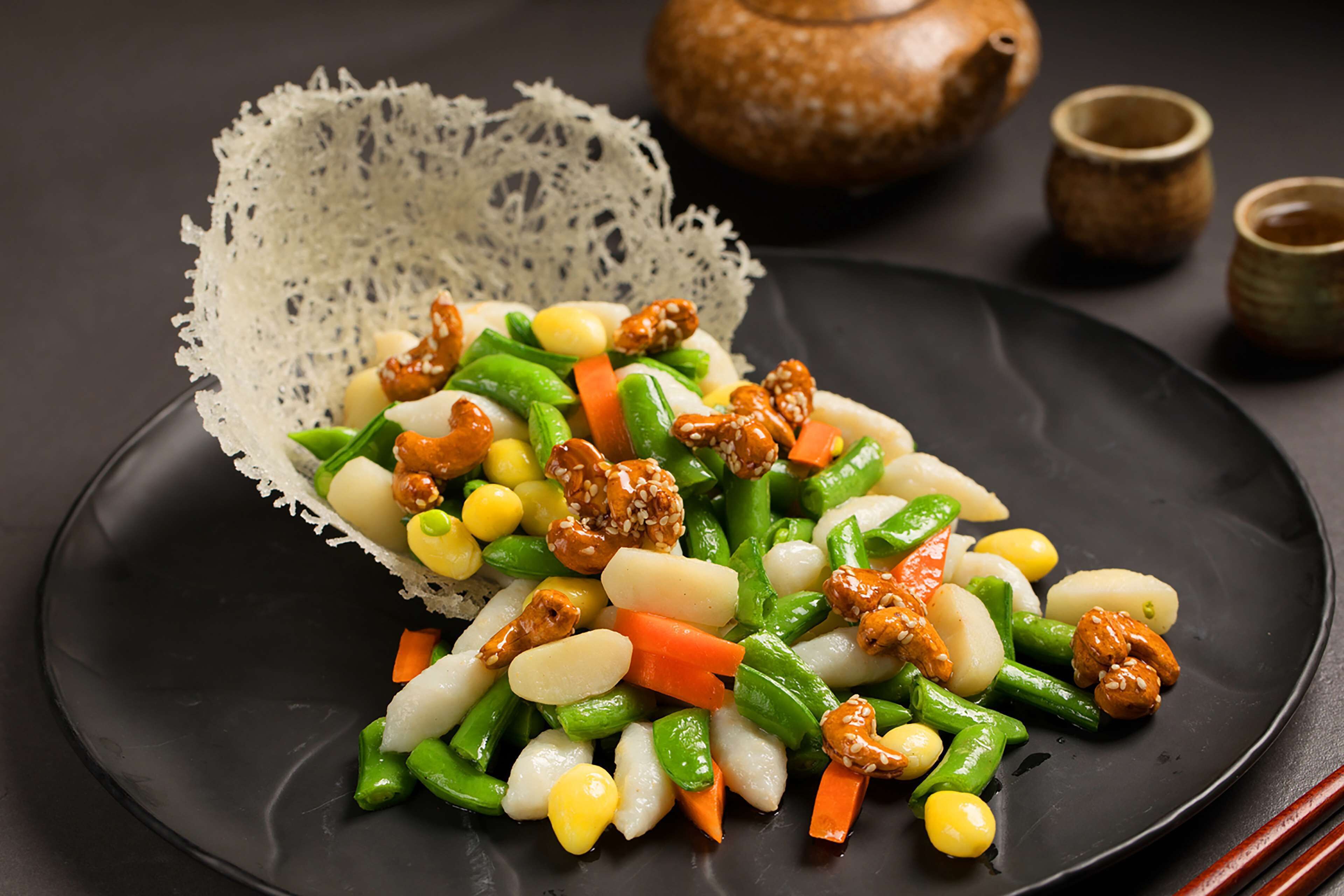 Un plato negro con una variedad de verduras y frutos secos, acompañado de un encaje blanco y una maceta marrón al fondo.
