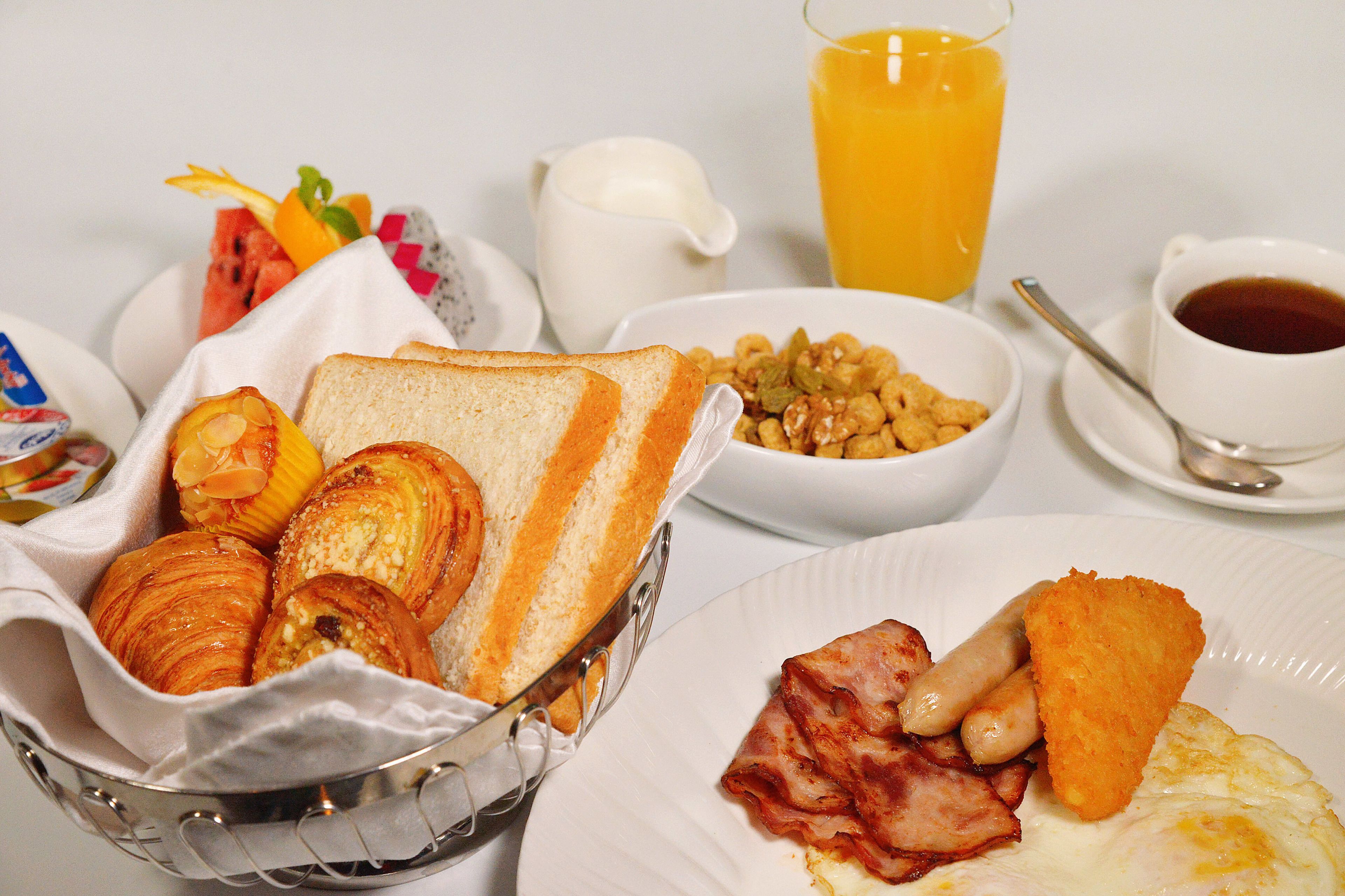 Une table de petit-déjeuner avec des croissants, des toasts, du bacon, des saucisses et un verre de jus d’orange.