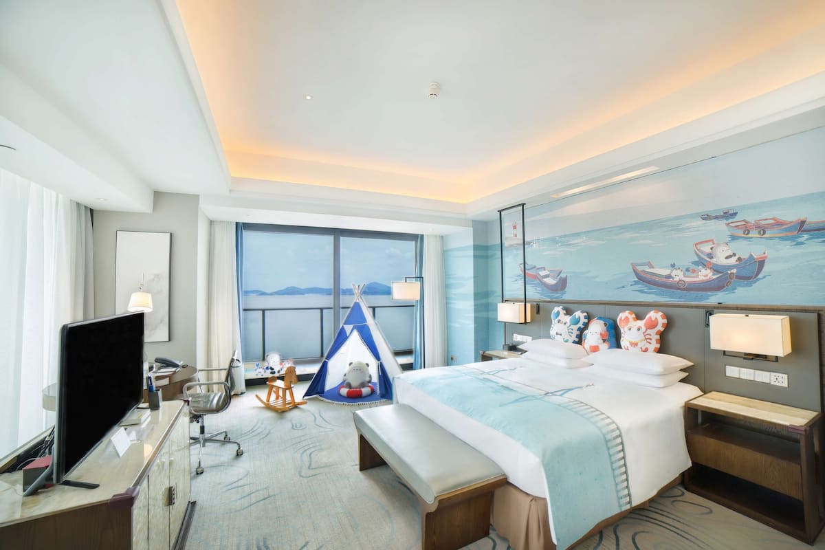 A hotel room with a large bed, a desk with a computer, a chair, a small table, a window with a view of the sea, and a painting of boats on the wall.