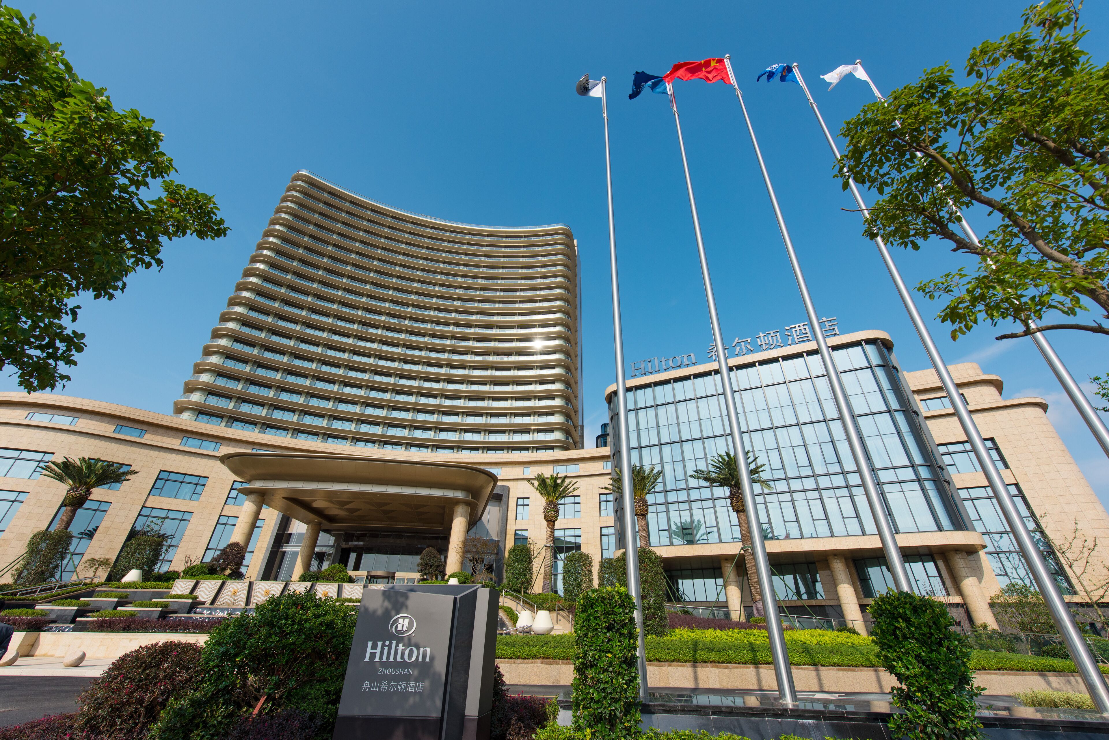 Un hotel moderno con fachada curva, varios mástiles de bandera y un jardín cuidado.