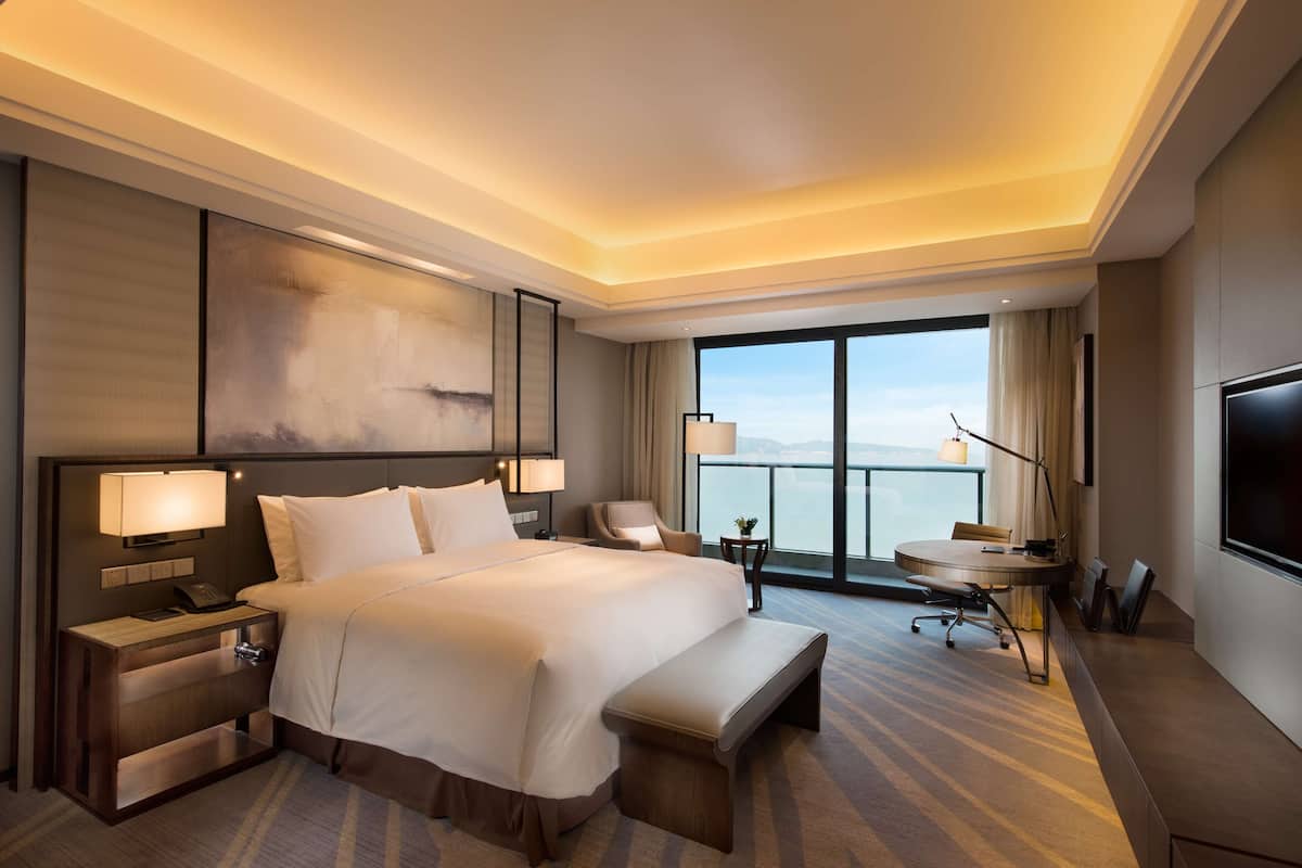 A hotel room with a large bed, bedside tables, a desk, a chair, a bench, and a large window with a view.