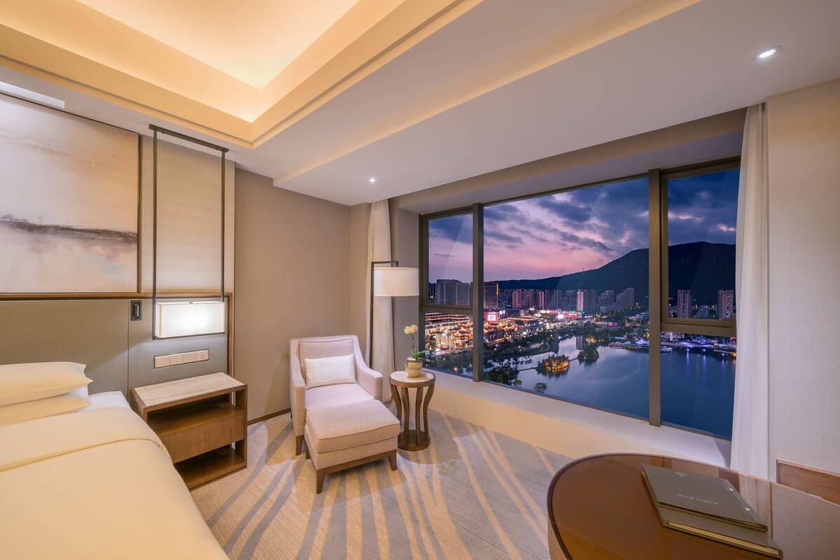 A hotel room with a large window offering a city view at dusk, a bed, a chair, a small table, and a desk with a laptop.