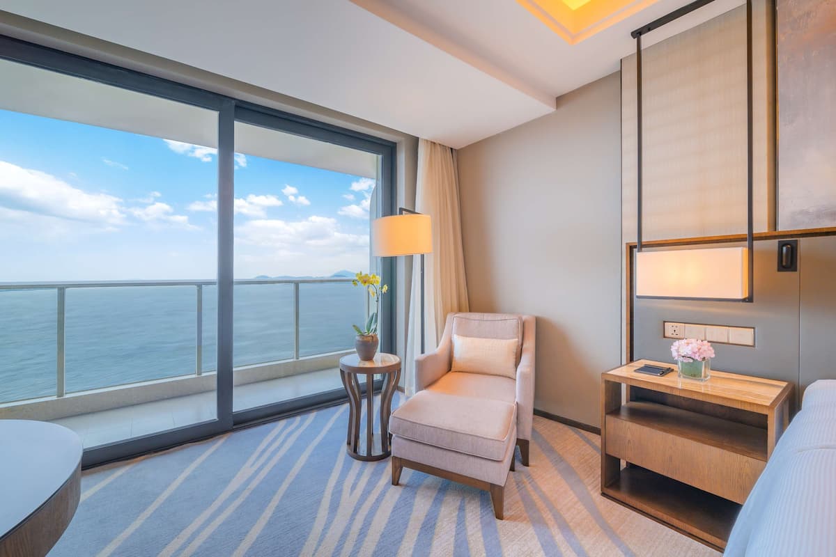 A hotel room with a large window offering a view of the sea, a comfortable chair, a small side table with a lamp, and a modern bed.