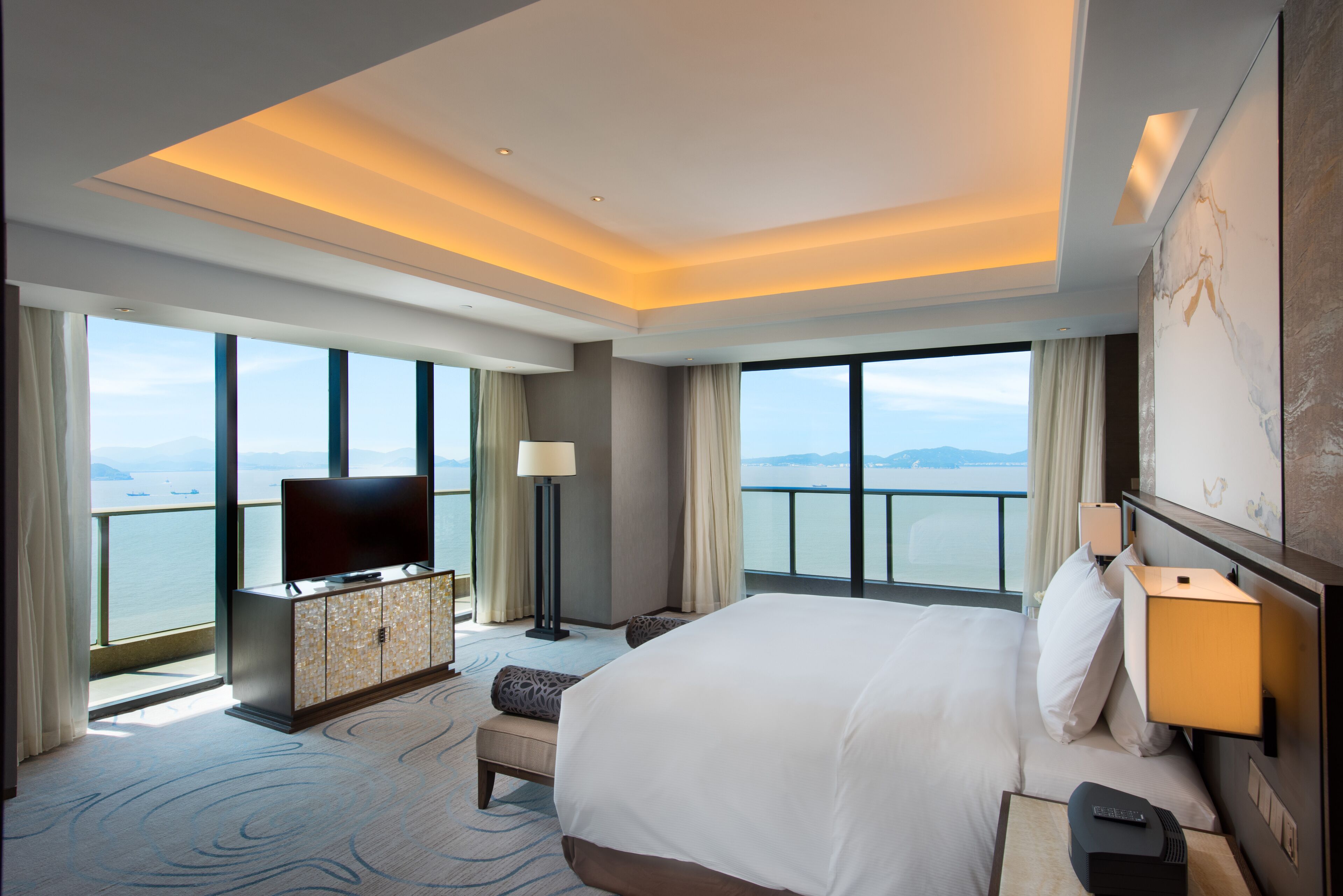 Habitación de hotel con una cama grande, un televisor de pantalla plana, un escritorio con lámpara y vistas al mar a través de amplias ventanas.