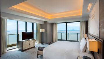 A hotel room with a large bed, a flat-screen TV, a desk with a lamp, and a view of the sea through large windows.
