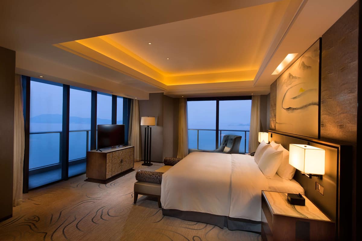 A hotel room with a large bed, a TV, a desk, and a view of the sea.