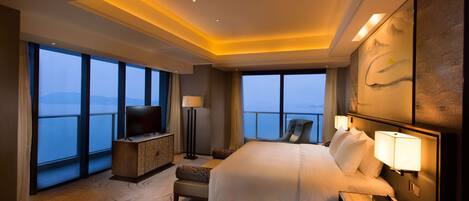 A hotel room with a large bed, a TV, a desk, and a view of the sea.