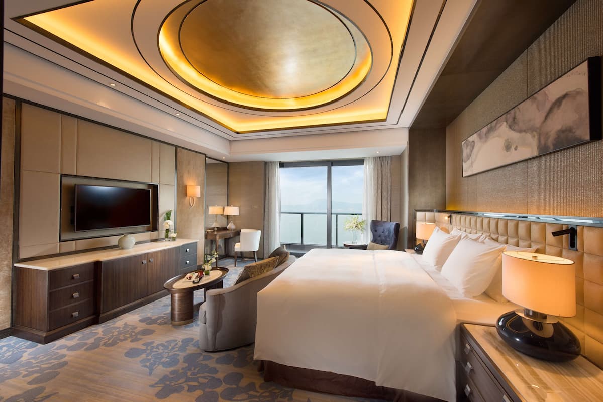 A modern hotel room with a large bed, a flat-screen TV, a seating area, and a view of the outdoors.