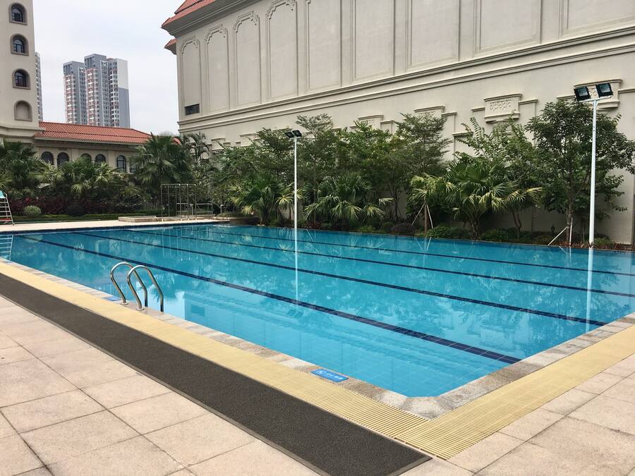A swimming pool with clear blue water, surrounded by a tiled deck and a building with classical architecture in the background.
