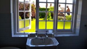 A bathroom with a window, a sink, and a view of a garden and houses. - The Queen Hotel (Olney)