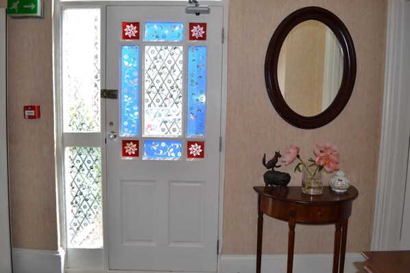 A room with a white door featuring decorative glass panels, a round mirror on the wall, and a small table with a vase of flowers. - The Queen Hotel (Olney)