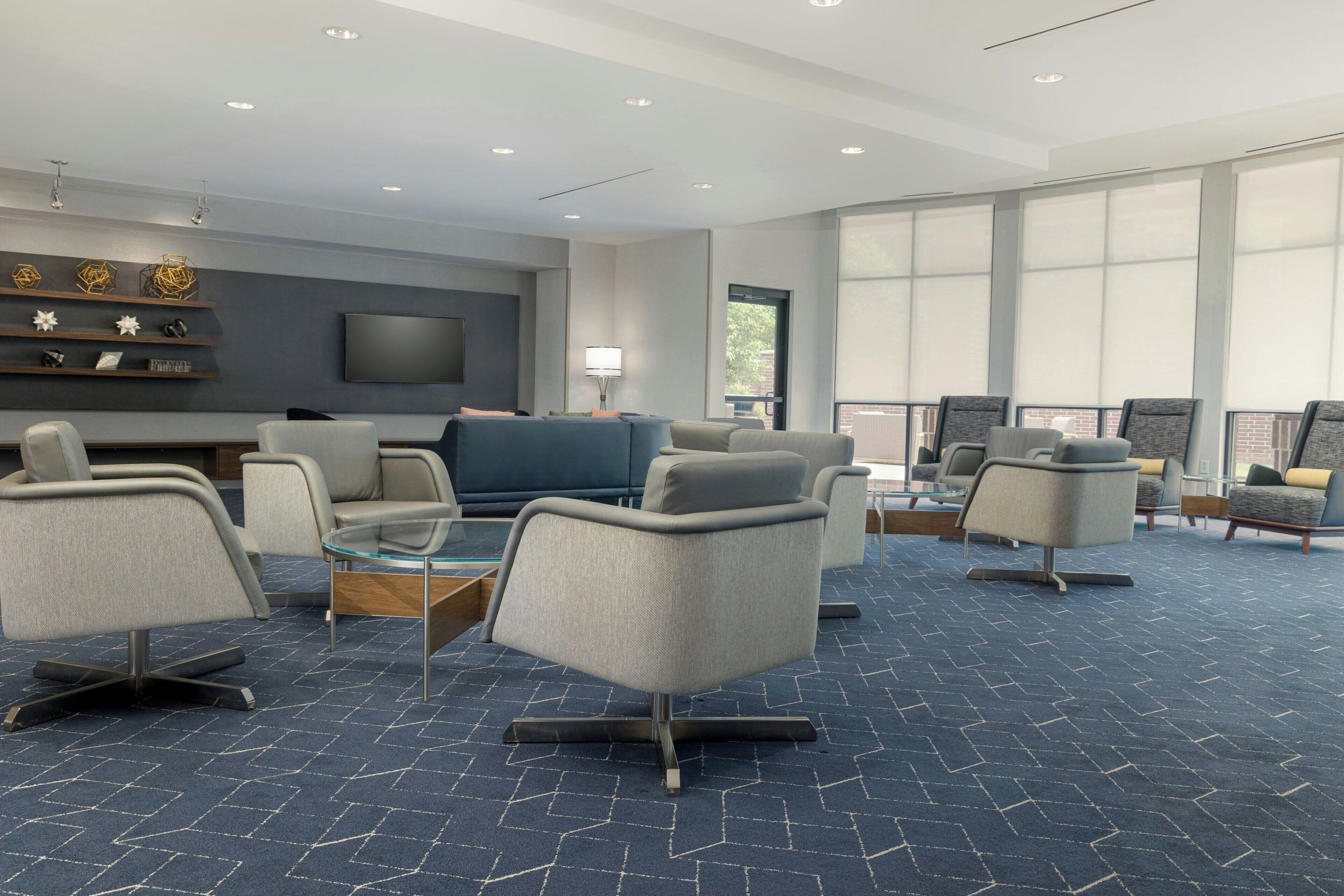 A modern hotel lobby with a blue carpet, grey upholstered chairs, a flat-screen TV, and a large window.