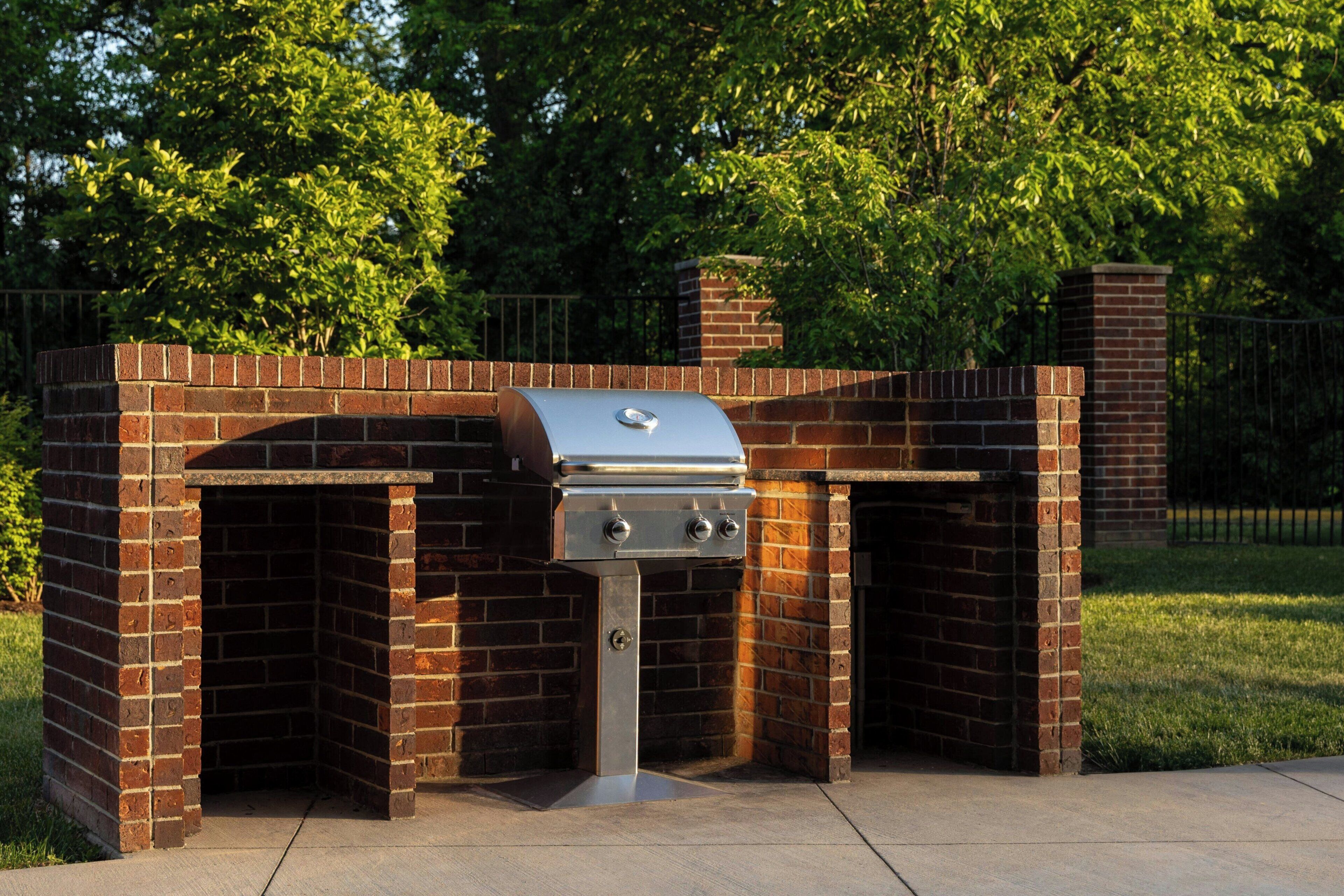 An outdoor brick barbecue grill with a chimney, surrounded by a brick wall and a metal fence.
