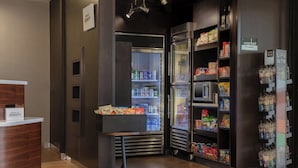 A hotel lobby with a reception desk, a refrigerator stocked with beverages, and shelves with snacks and bottled water.