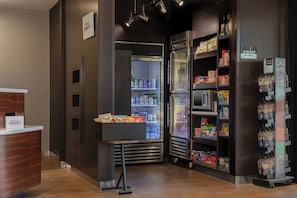 A hotel lobby with a reception desk, a refrigerator stocked with beverages, and shelves with snacks and bottled water.