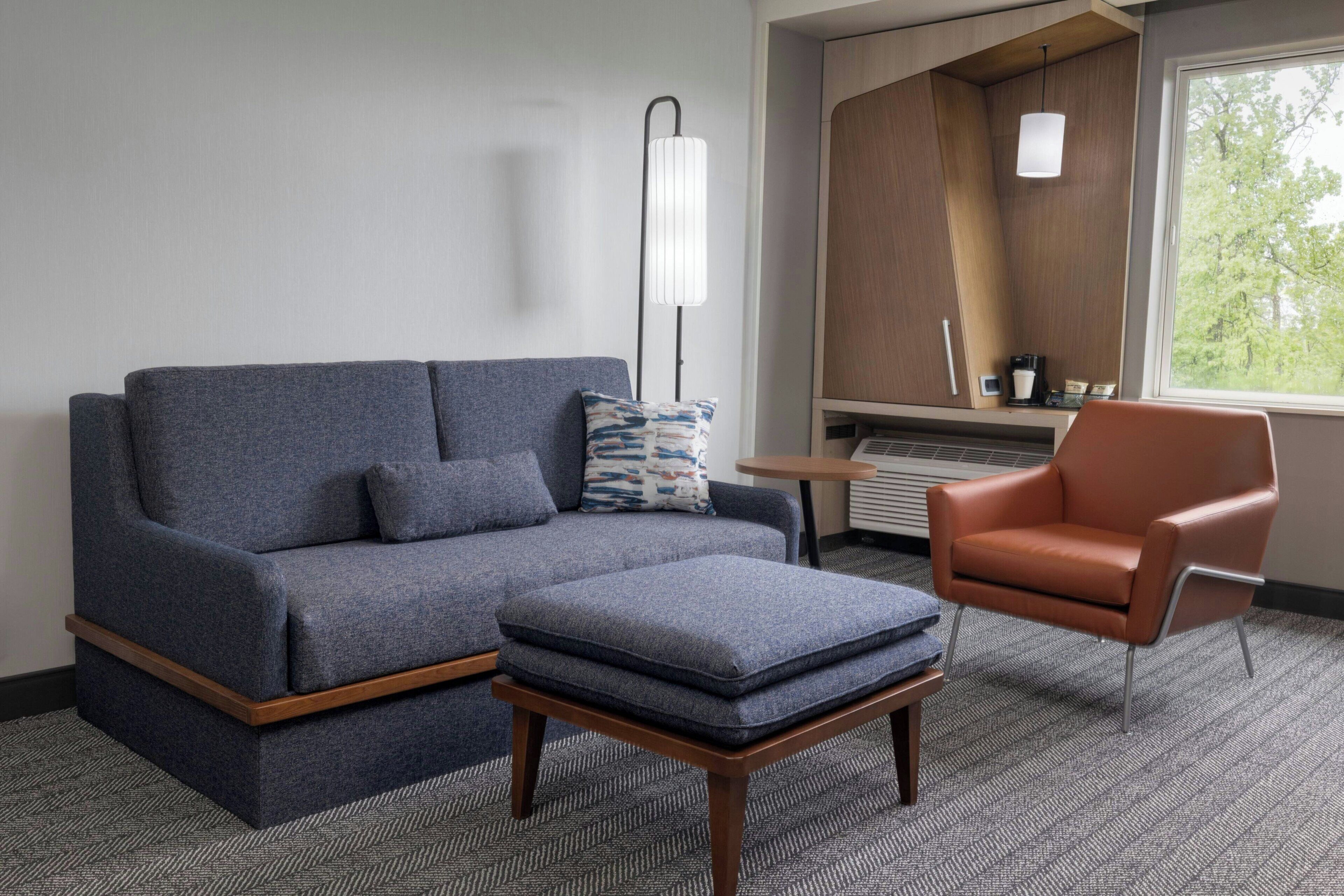 Una habitación de hotel moderna con un sofá gris, un pequeño reposapiés, un sillón de cuero marrón, una lámpara de pie y vistas al exterior a través de una ventana.