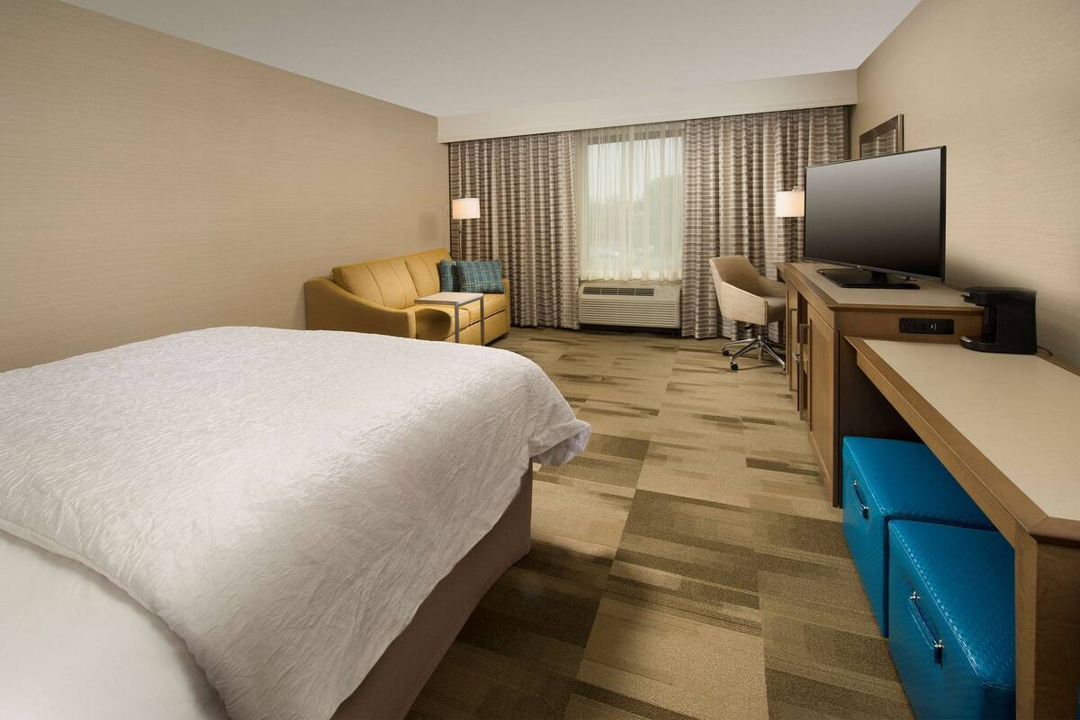 A hotel room with a large bed, a desk with a television, a chair, and a desk lamp.
