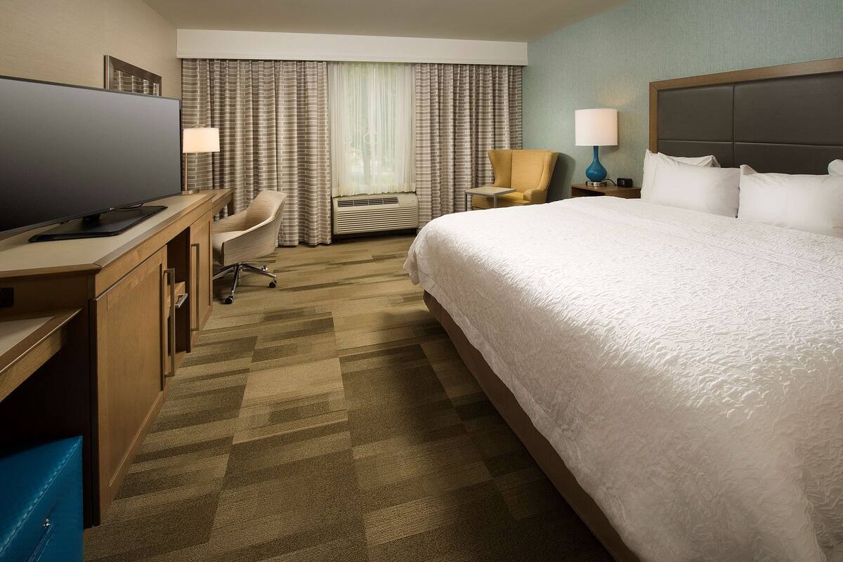 A hotel room with a large bed, a desk with a flat-screen TV, a chair, an air conditioning unit, and a nightstand with a lamp.