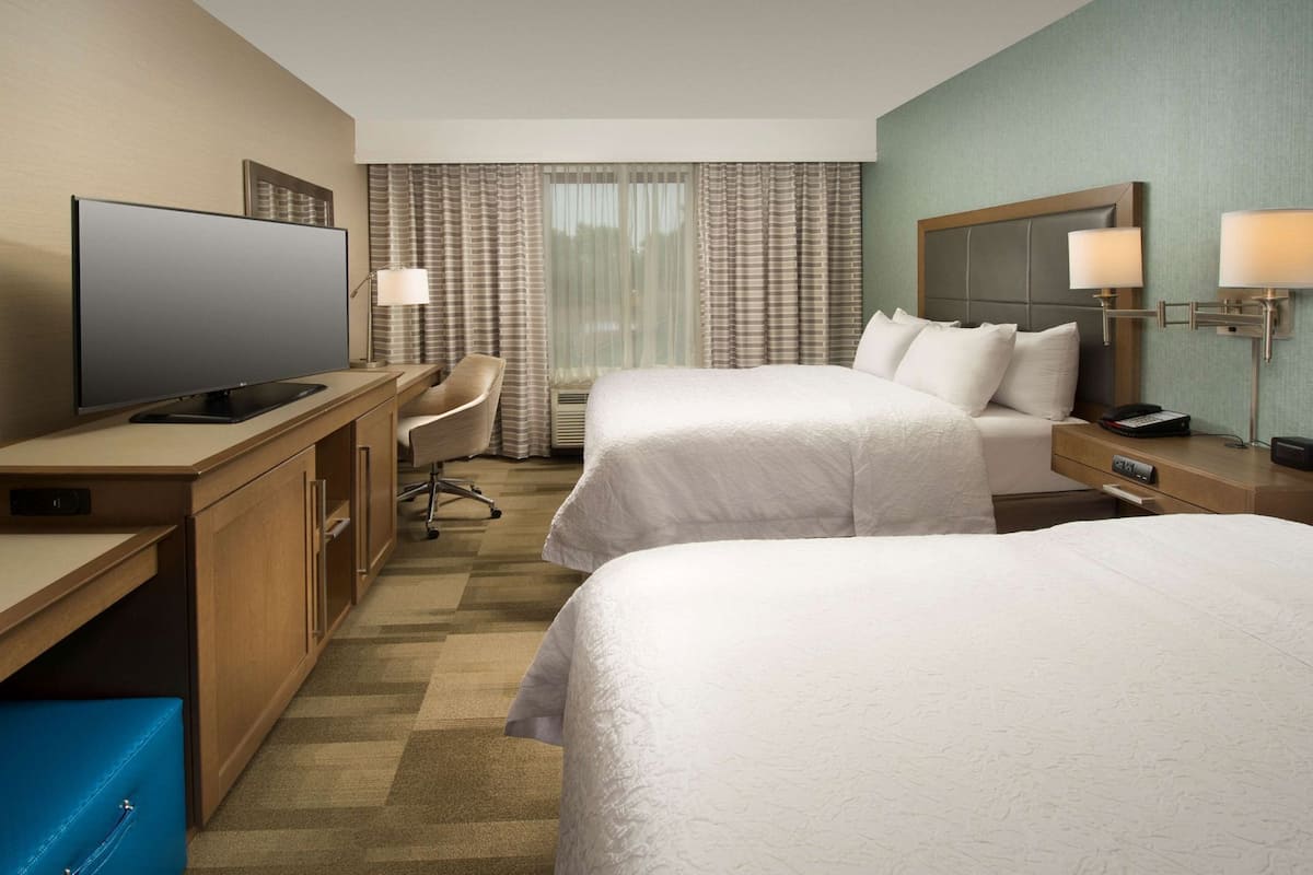 A hotel room with two beds, a desk with a television, a chair, and a desk lamp.