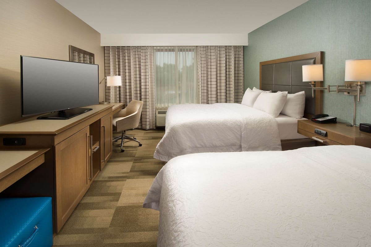 A hotel room with two beds, a desk with a flat-screen TV, a chair, and a desk lamp.