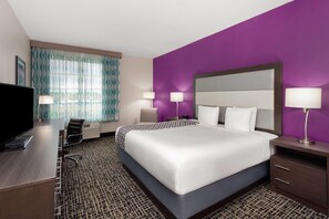 A hotel room with a large bed, a desk with a computer, two bedside lamps, and a window with patterned curtains.