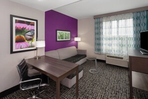Une chambre d’hôtel équipée d’une table à manger, d’un canapé, d’une télévision, d’une lampe et d’une fenêtre avec des rideaux à motifs.
