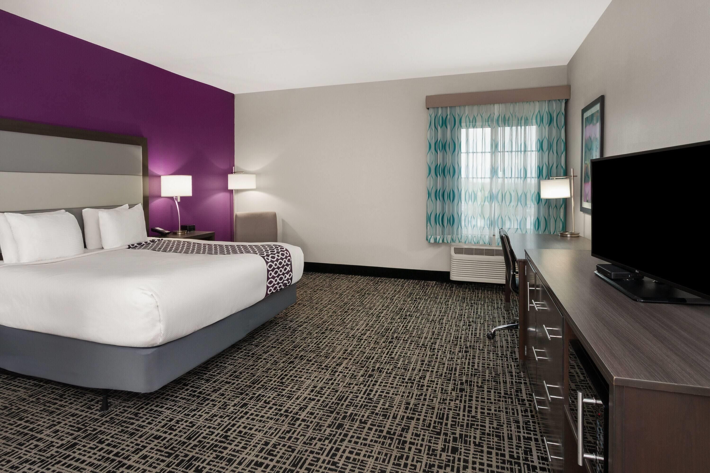 Habitación de hotel con cama, escritorio, televisor, ventana con cortinas y alfombra estampada.