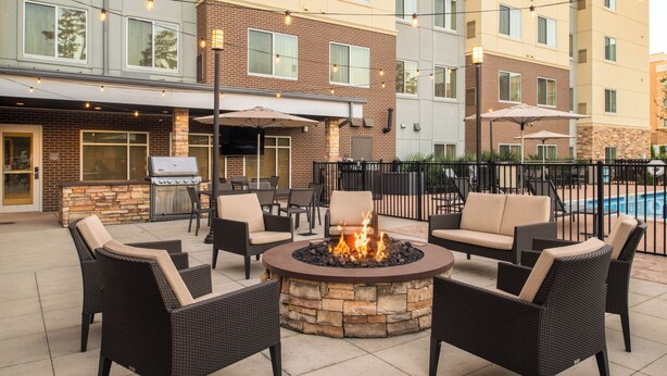 A patio area with a stone fire pit surrounded by wicker chairs, a grill, and umbrellas. - Residence Inn by Marriott Houston City Place (Spring)