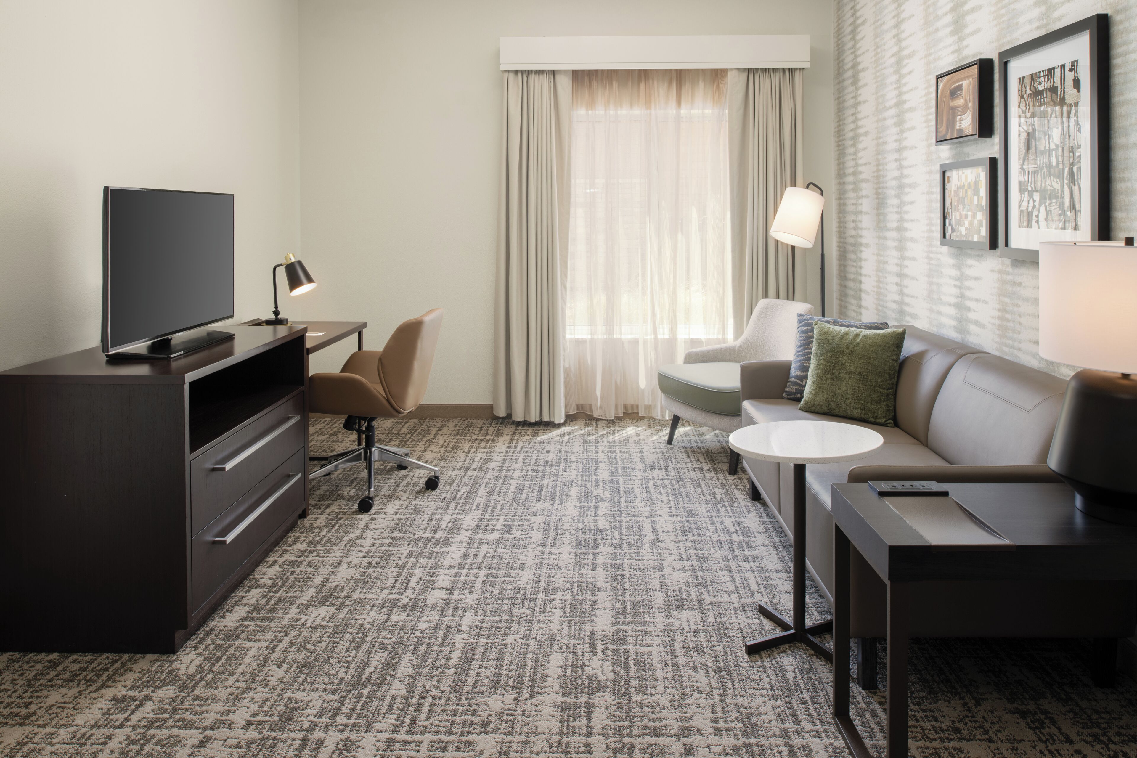 A hotel room with a flat-screen TV, a desk with a lamp, a sofa with a remote, and a patterned carpet.