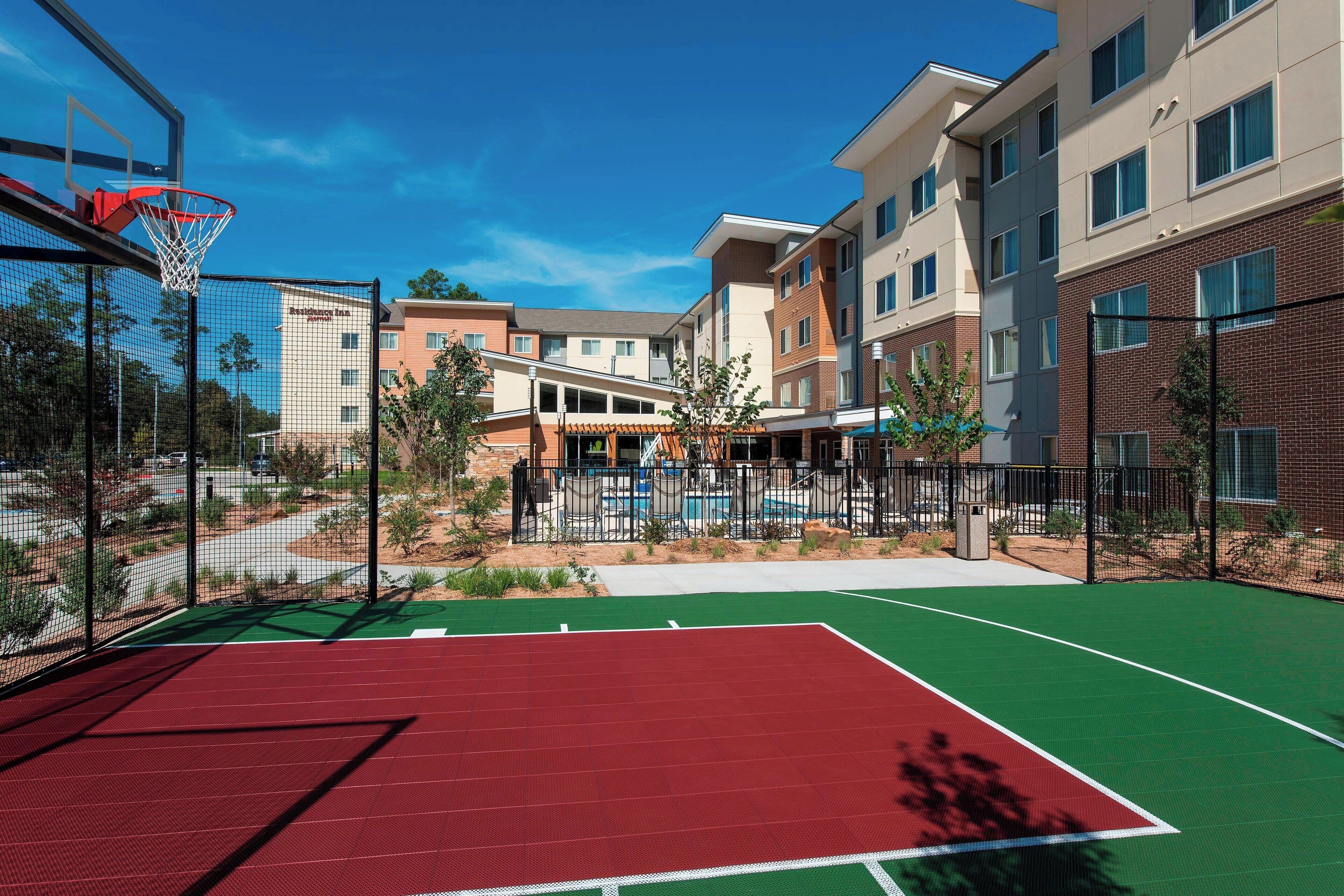 Un terrain de basket avec un panier, entouré d’une clôture, et situé à côté d’un bâtiment doté de nombreuses fenêtres et d’un espace abrité.