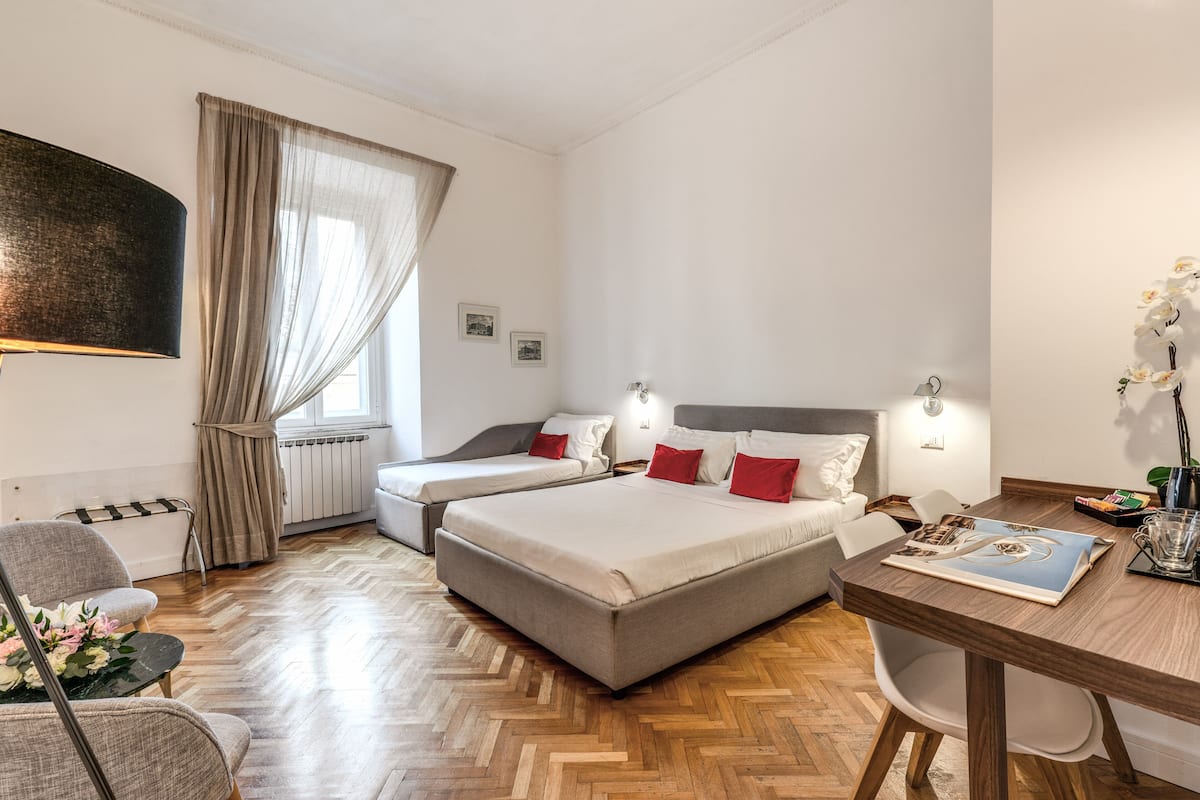 A hotel room with two beds, a dining table, a chair, a radiator, and a window with curtains.