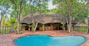 A thatched-roof building with a pool and outdoor seating area surrounded by trees.