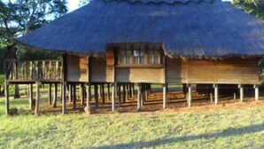 Una estructura con techo de paja y fachada de madera, sostenida por pilotes, en un área cubierta de pasto.