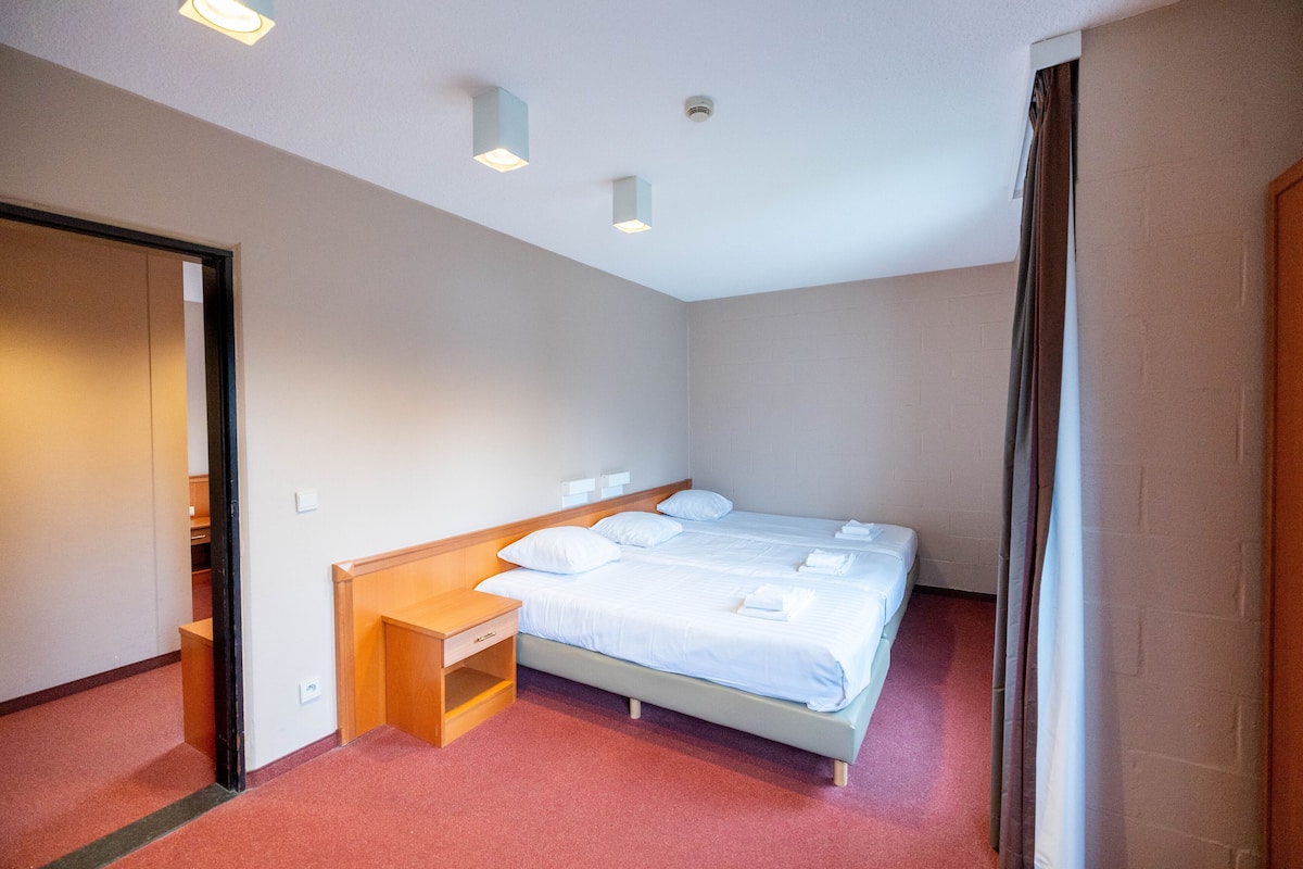 Une chambre d’hôtel avec un lit simple, une table de chevet en bois, un petit bureau et une armoire.