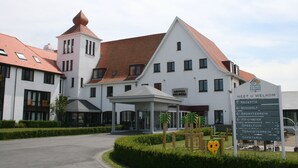 Een groot wit gebouw met een rood dak, een kleine toren en een bord waarop faciliteiten staan aangegeven, zoals receptie, fitnessruimte en cafetaria.