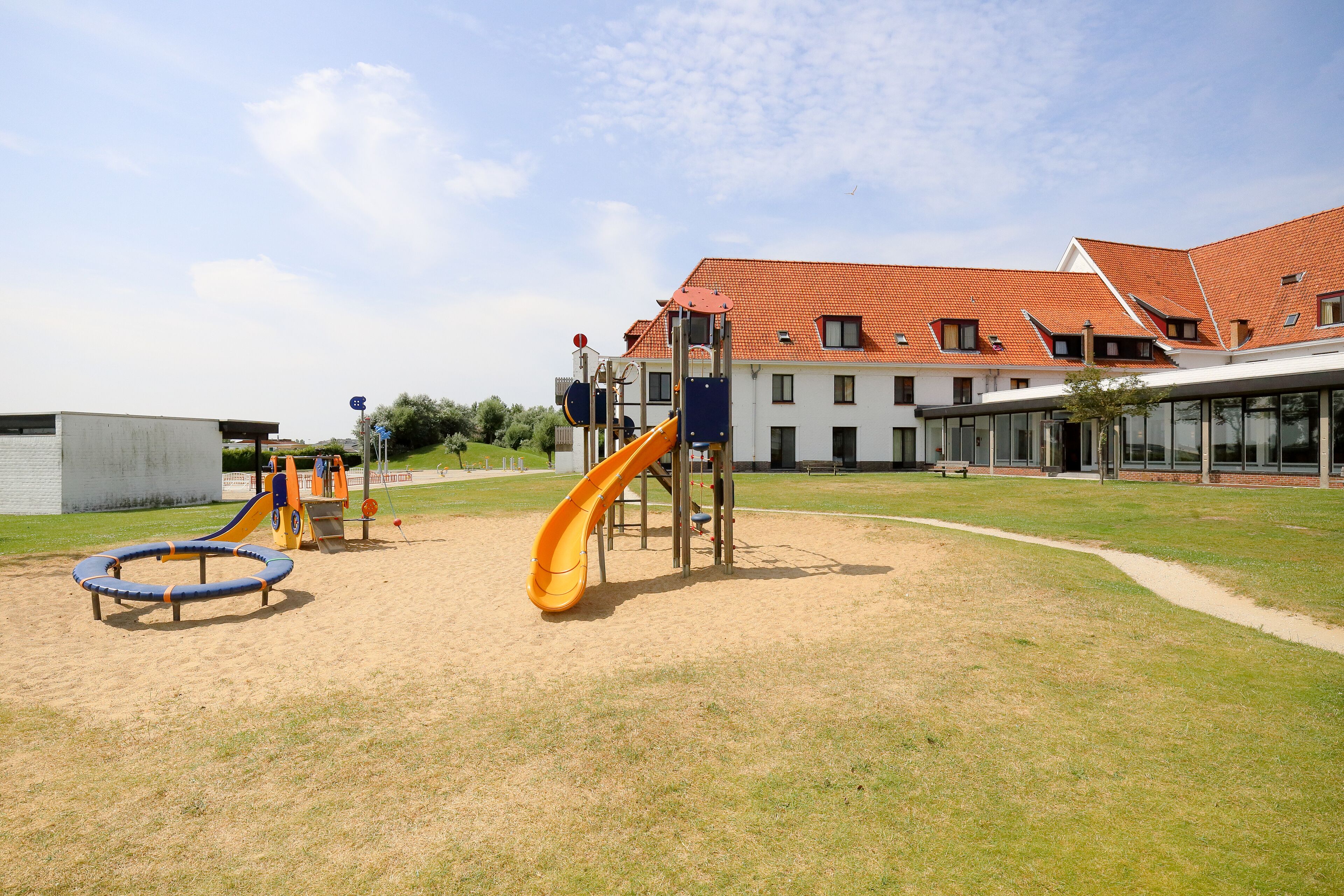 A playground with a slide and swings, surrounded by a grassy area and a building with a red-tiled roof.